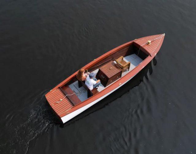 A small wooden motorboat with two people seated, moving across calm dark water.