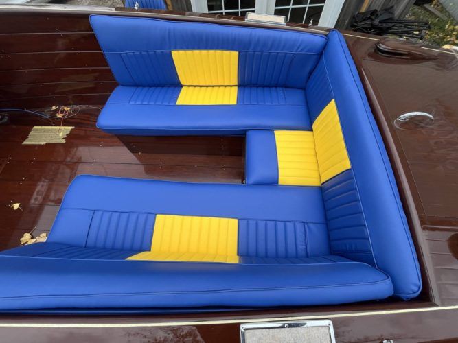 Interior of a classic wooden boat featuring blue and yellow upholstered seating and a vintage steering wheel inside a workshop.