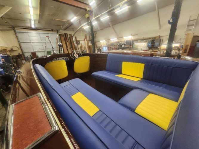 Interior of a classic wooden boat featuring blue and yellow upholstered seating and a vintage steering wheel inside a workshop.