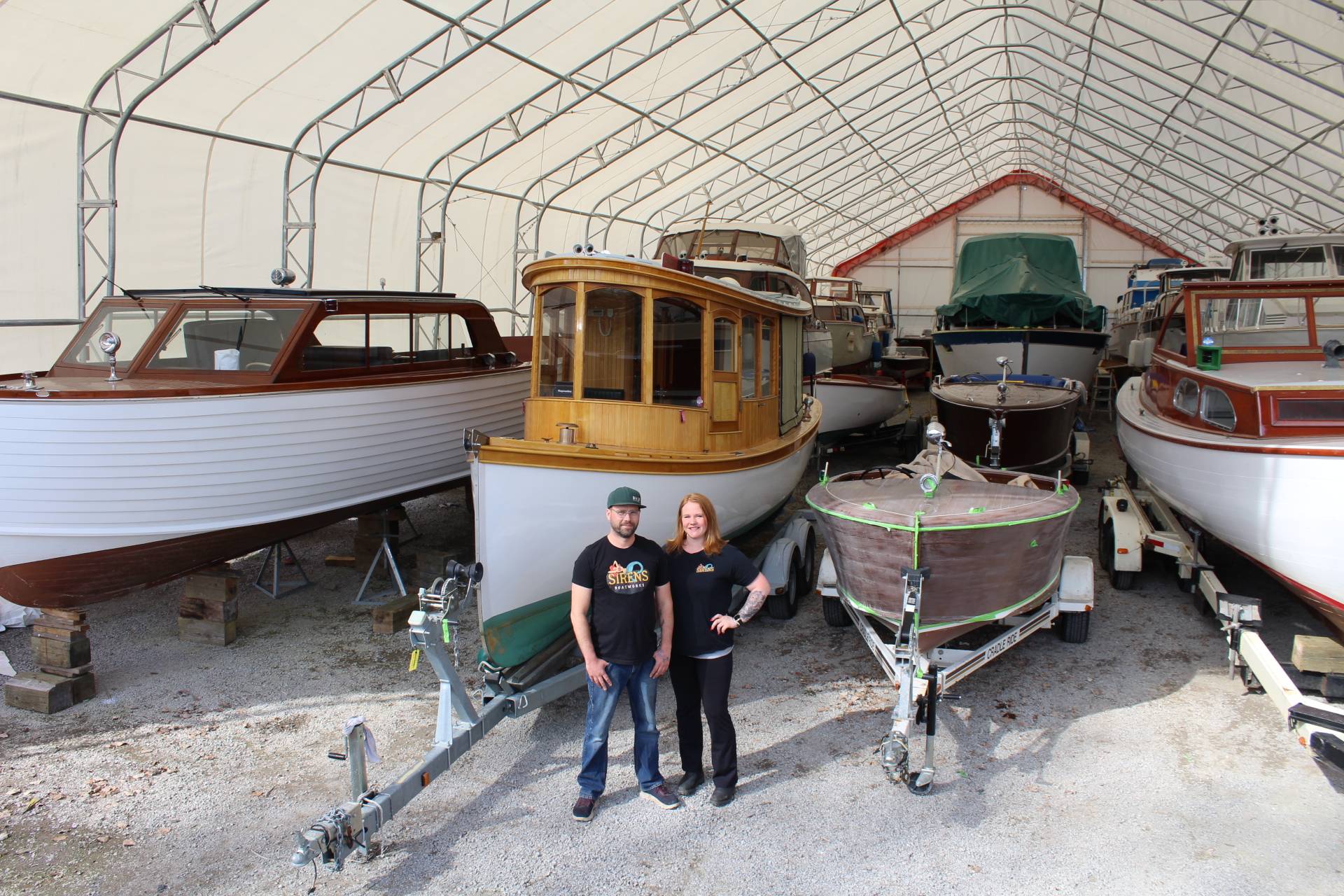 Two people standing among classic wooden boats stored inside a large covered boat facility.